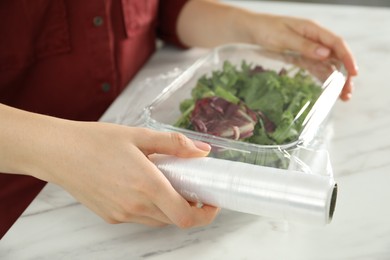 Photo of Woman putting plastic food wrap over glass container with salad at white table in kitchen, closeup