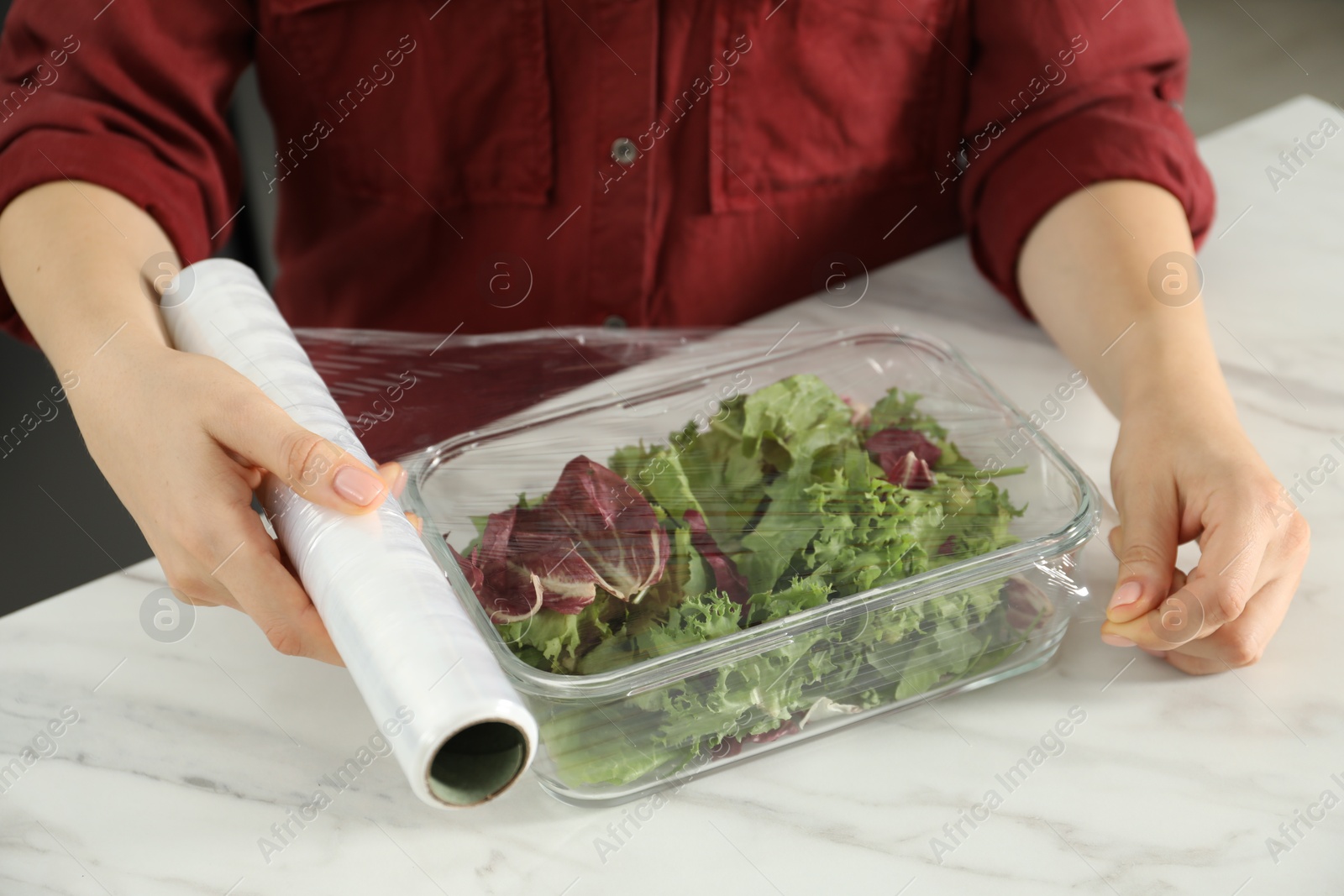 Photo of Woman putting plastic food wrap over glass container with salad at white table in kitchen, closeup