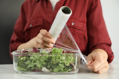 Photo of Woman putting plastic food wrap over glass container with salad at white table in kitchen, closeup