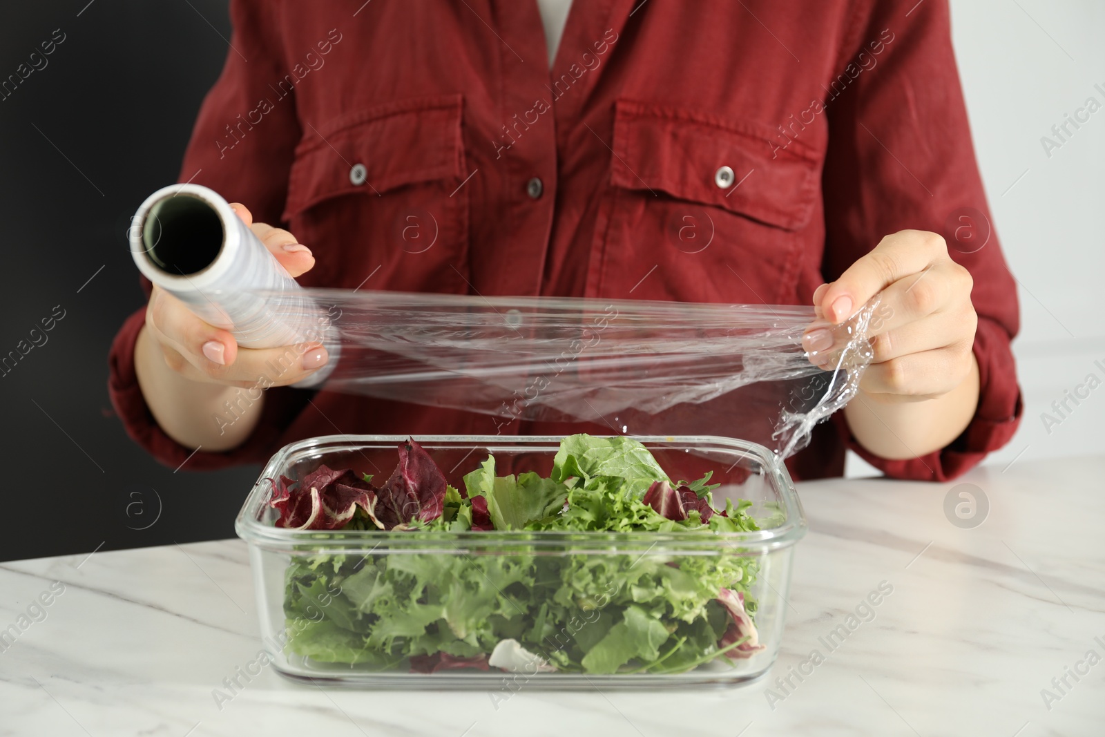 Photo of Woman putting plastic food wrap over glass container with salad at white table in kitchen, closeup