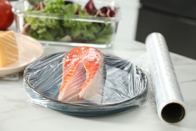 Plate of salmon with plastic food wrap on white marble table in kitchen, closeup