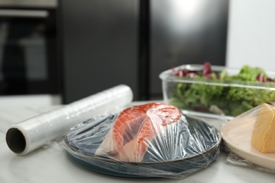 Plate of salmon with plastic food wrap on white table in kitchen, closeup