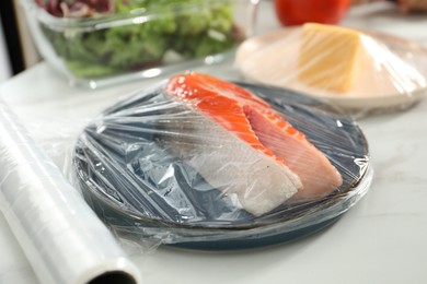 Plate of salmon with plastic food wrap on white table in kitchen, closeup