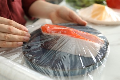 Woman putting plastic food wrap over plate with salmon at white table in kitchen, closeup