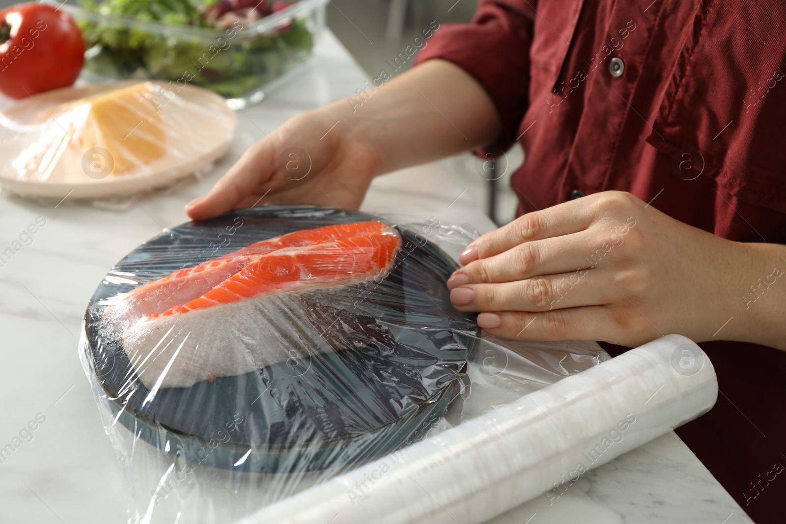 Photo of Woman putting plastic food wrap over plate with salmon at white marble table in kitchen, closeup