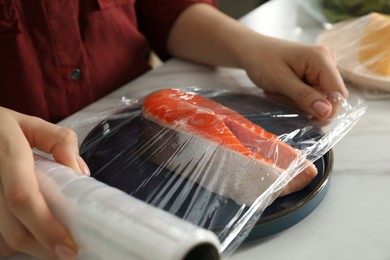 Woman putting plastic food wrap over plate with salmon at white marble table in kitchen, closeup