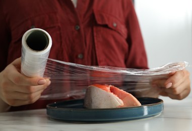 Woman putting plastic food wrap over plate with salmon at white marble table in kitchen, closeup