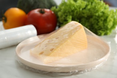 Photo of Plate of cheese with plastic food wrap and other products on white table, closeup