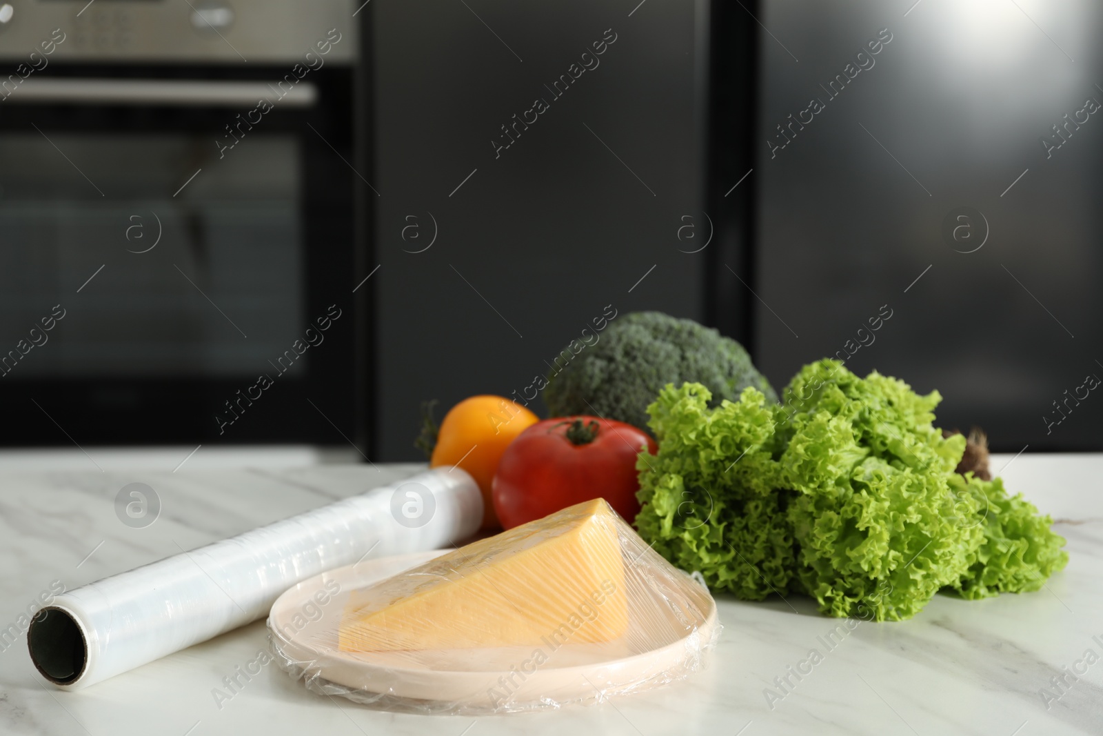 Photo of Plate of cheese with plastic food wrap and other products on white marble table in kitchen