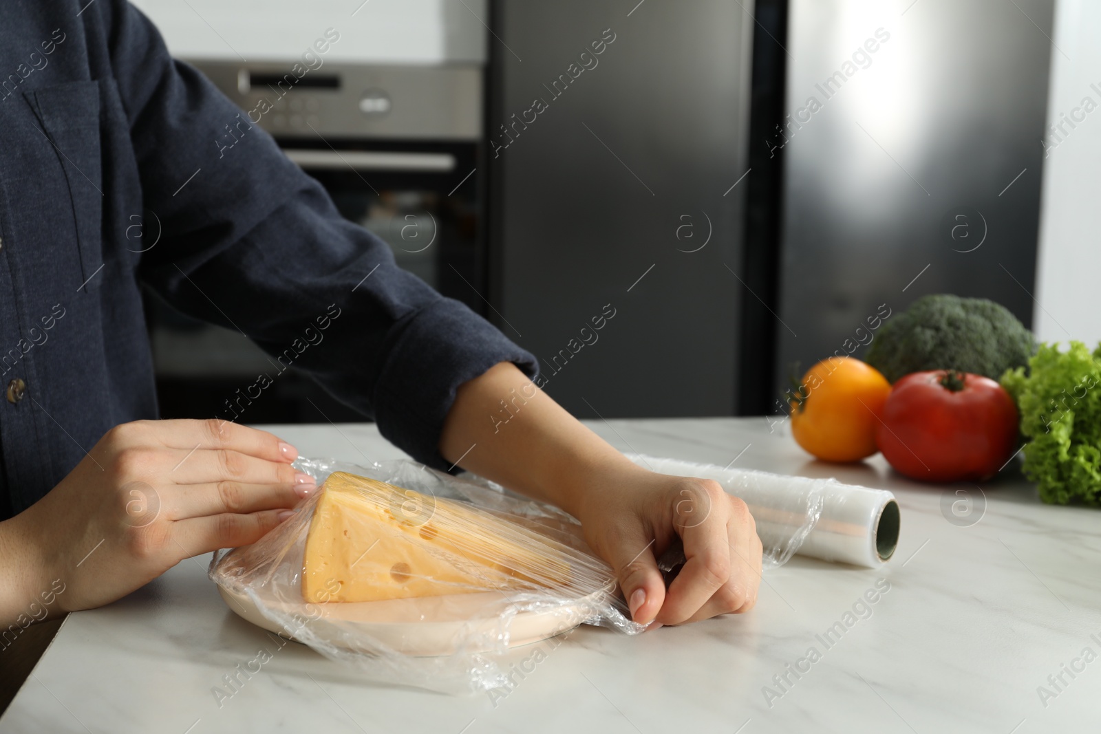 Photo of Woman putting plastic food wrap over plate with cheese at white marble table in kitchen, closeup