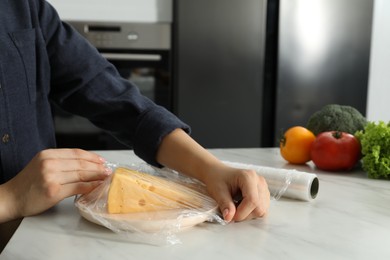 Woman putting plastic food wrap over plate with cheese at white marble table in kitchen, closeup