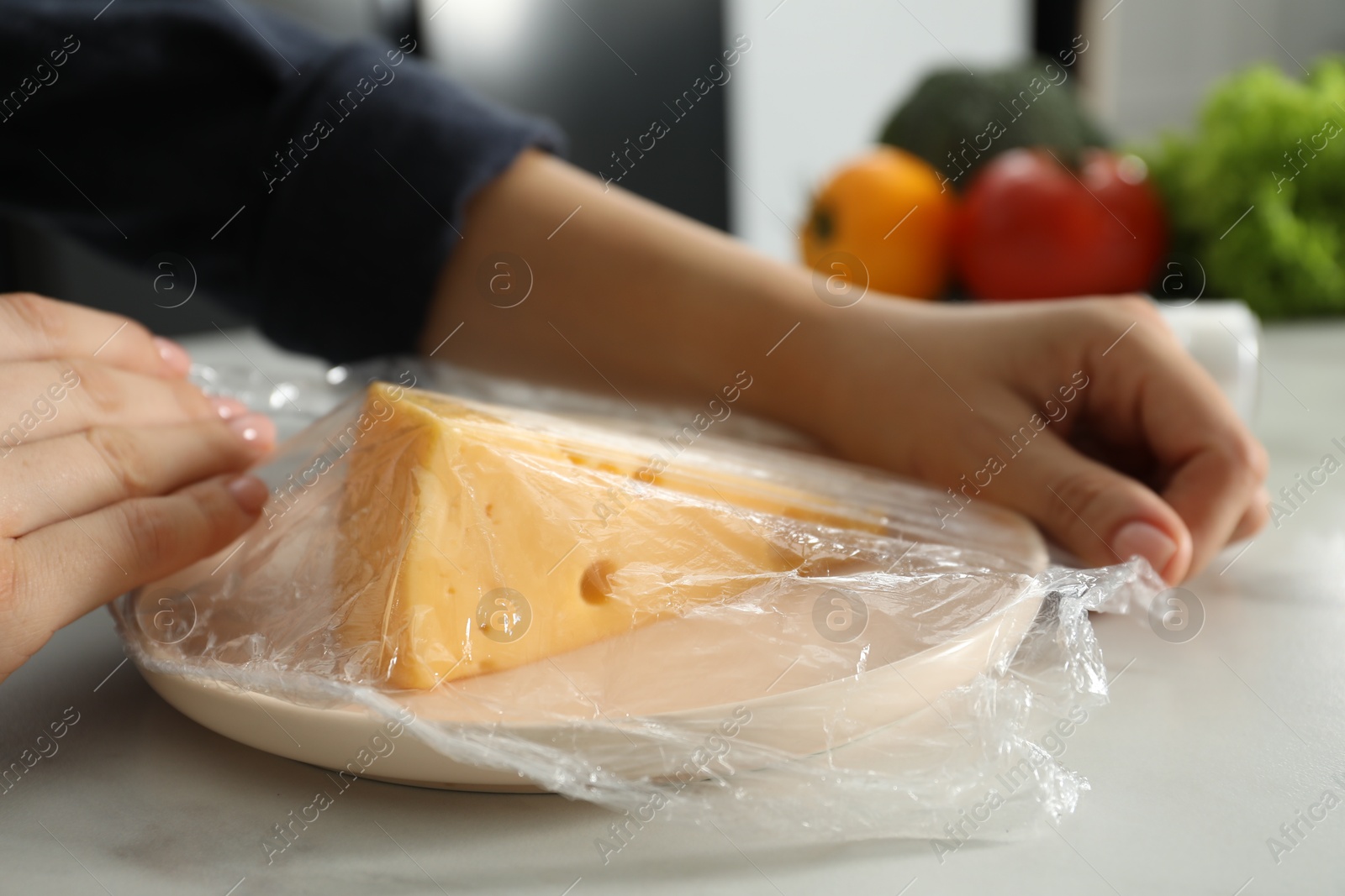 Photo of Woman putting plastic food wrap over plate with cheese at white marble table in kitchen, closeup