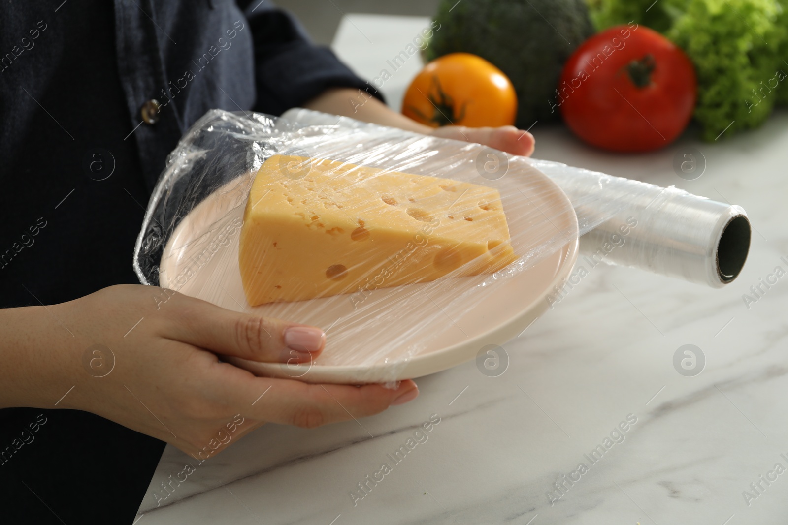 Photo of Woman putting plastic food wrap over plate with cheese at white marble table in kitchen, closeup