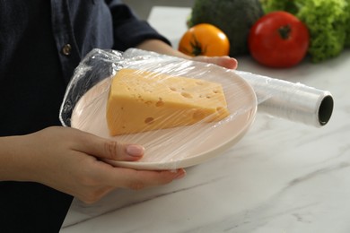 Woman putting plastic food wrap over plate with cheese at white marble table in kitchen, closeup