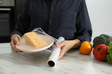 Woman putting plastic food wrap over plate with cheese at white marble table in kitchen, closeup