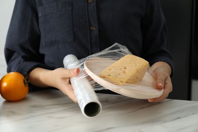 Photo of Woman putting plastic food wrap over plate with cheese at white marble table in kitchen, closeup