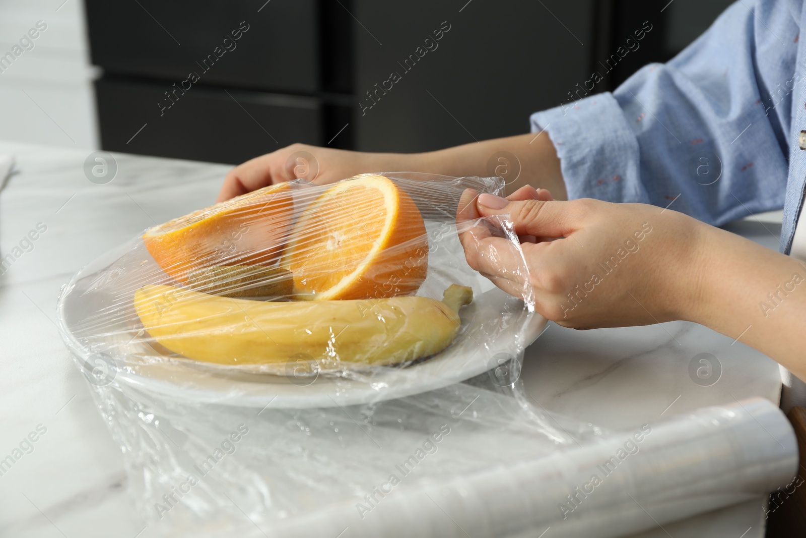 Photo of Woman putting plastic food wrap over plate with fruits at countertop in kitchen, closeup