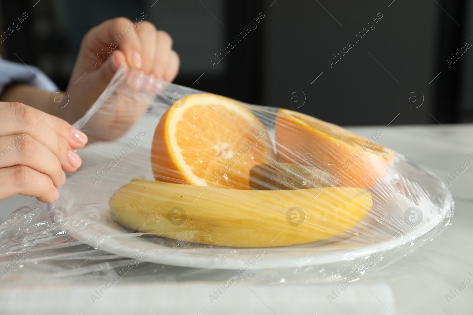 Photo of Woman putting plastic food wrap over plate with fruits at countertop in kitchen, closeup