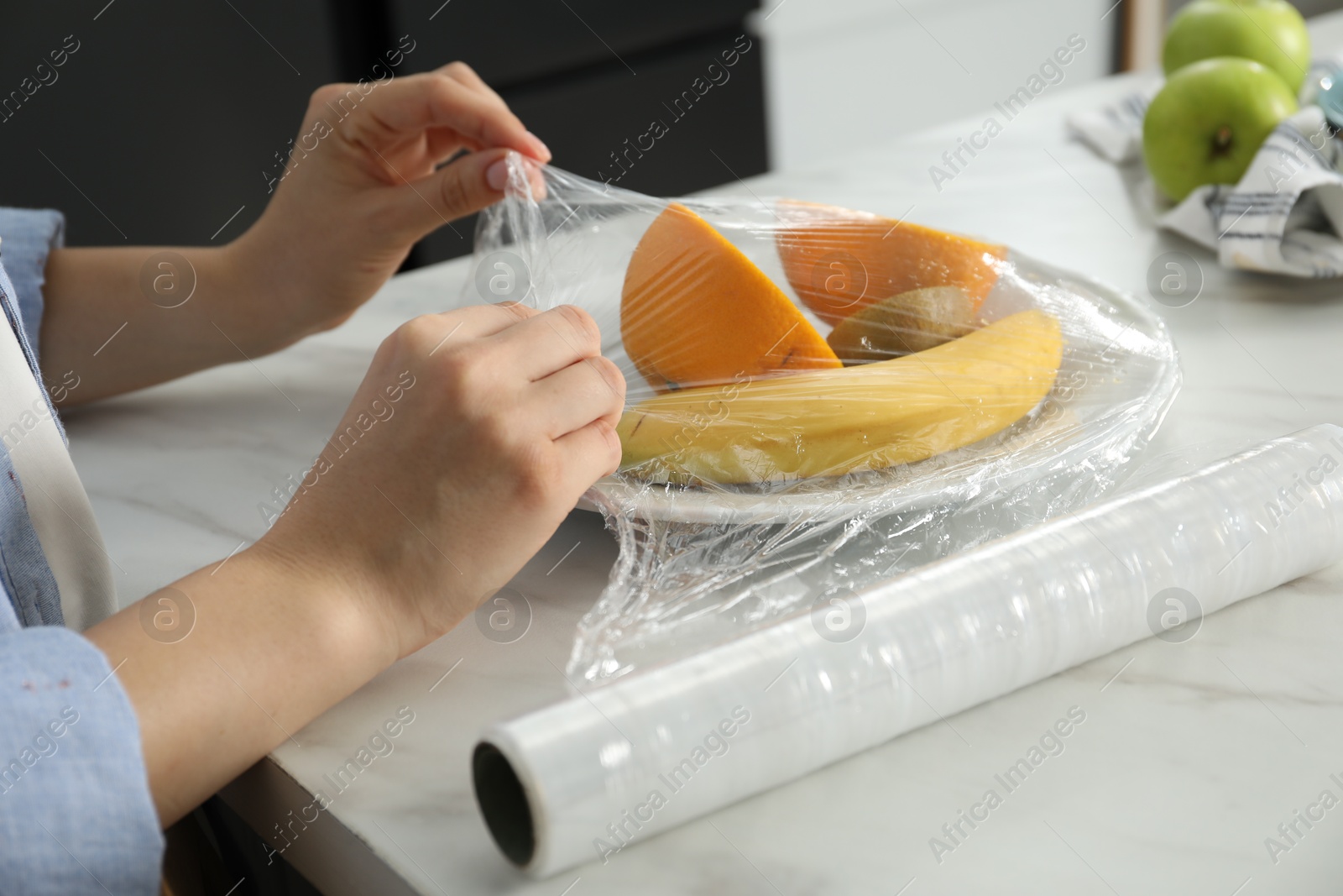 Photo of Woman putting plastic food wrap over plate with fruits at countertop in kitchen, closeup