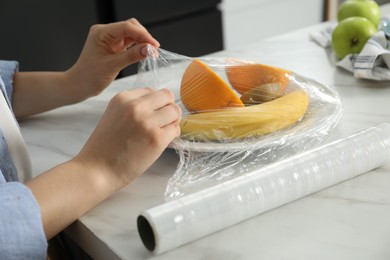 Woman putting plastic food wrap over plate with fruits at countertop in kitchen, closeup