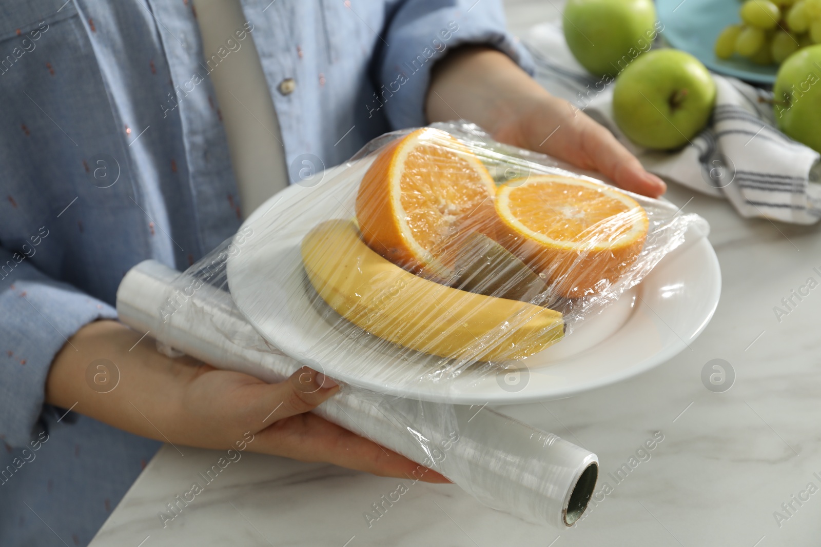 Photo of Woman putting plastic food wrap over plate with fruits at countertop in kitchen, closeup