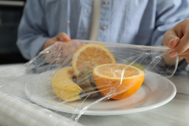 Woman putting plastic food wrap over plate with fruits at countertop in kitchen, closeup