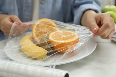 Woman putting plastic food wrap over plate with fruits at countertop in kitchen, closeup