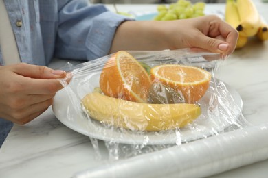 Woman putting plastic food wrap over plate with fruits at countertop in kitchen, closeup