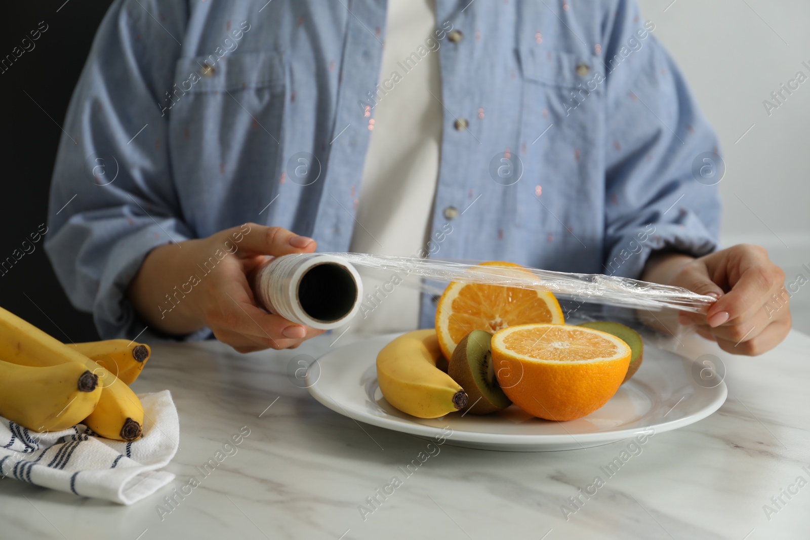 Photo of Woman putting plastic food wrap over plate with fruits at countertop in kitchen, closeup