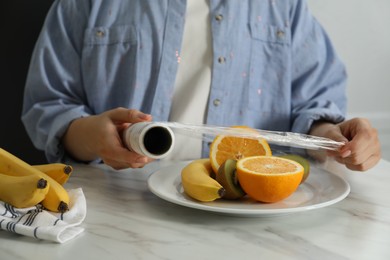 Photo of Woman putting plastic food wrap over plate with fruits at countertop in kitchen, closeup