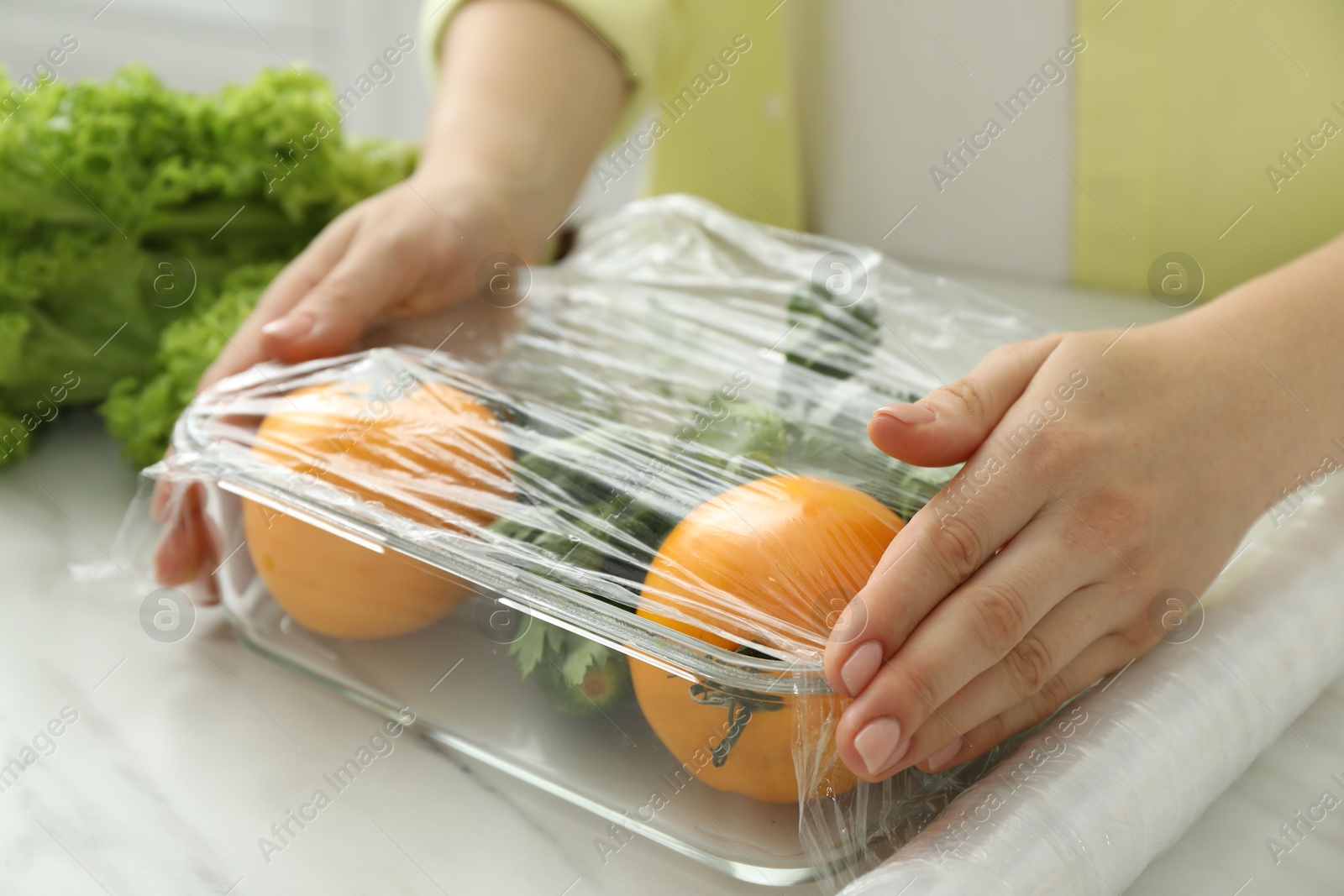 Photo of Woman putting plastic food wrap over glass container with fresh vegetables at white marble table, closeup