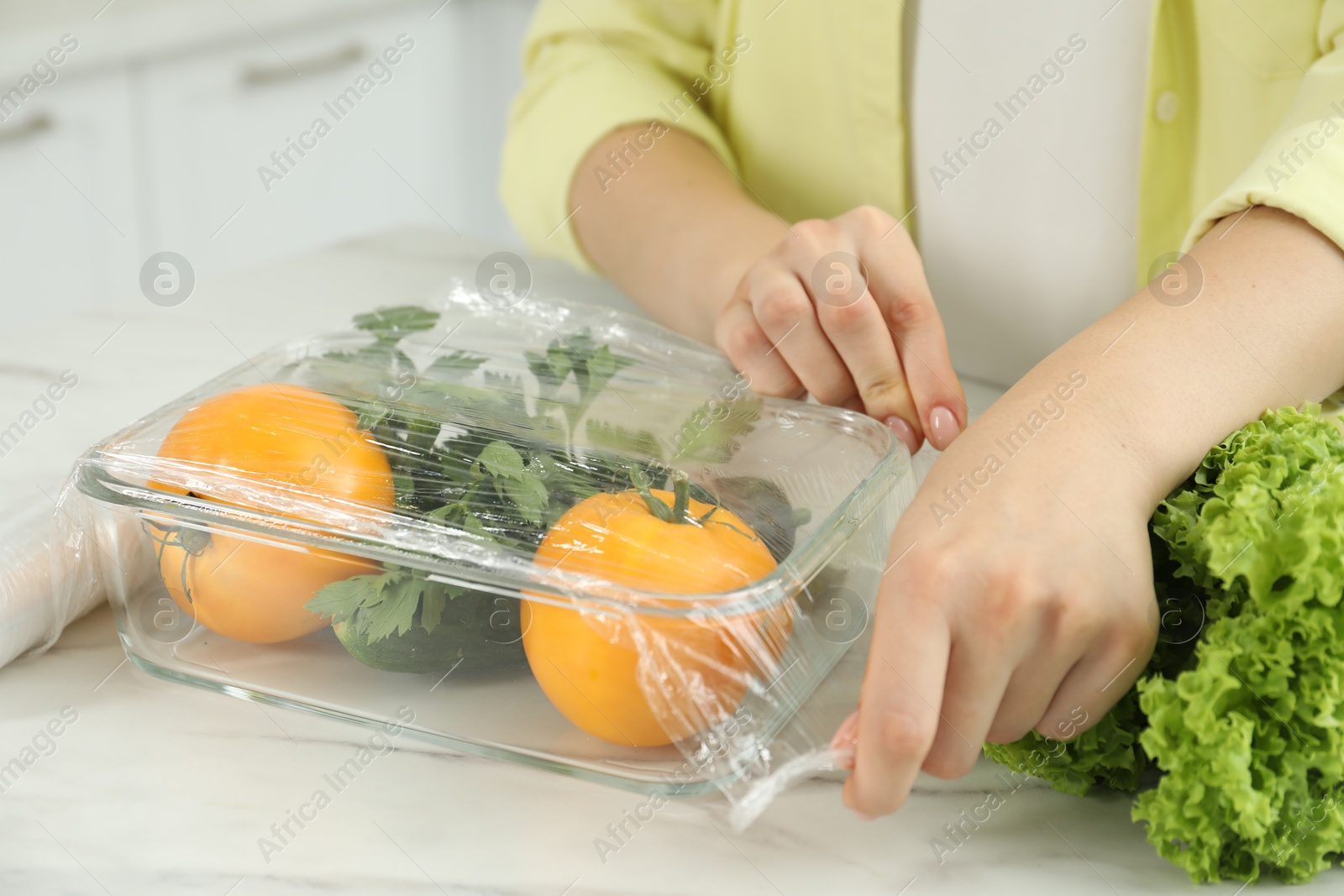 Photo of Woman putting plastic food wrap over glass container with fresh vegetables at white marble table, closeup