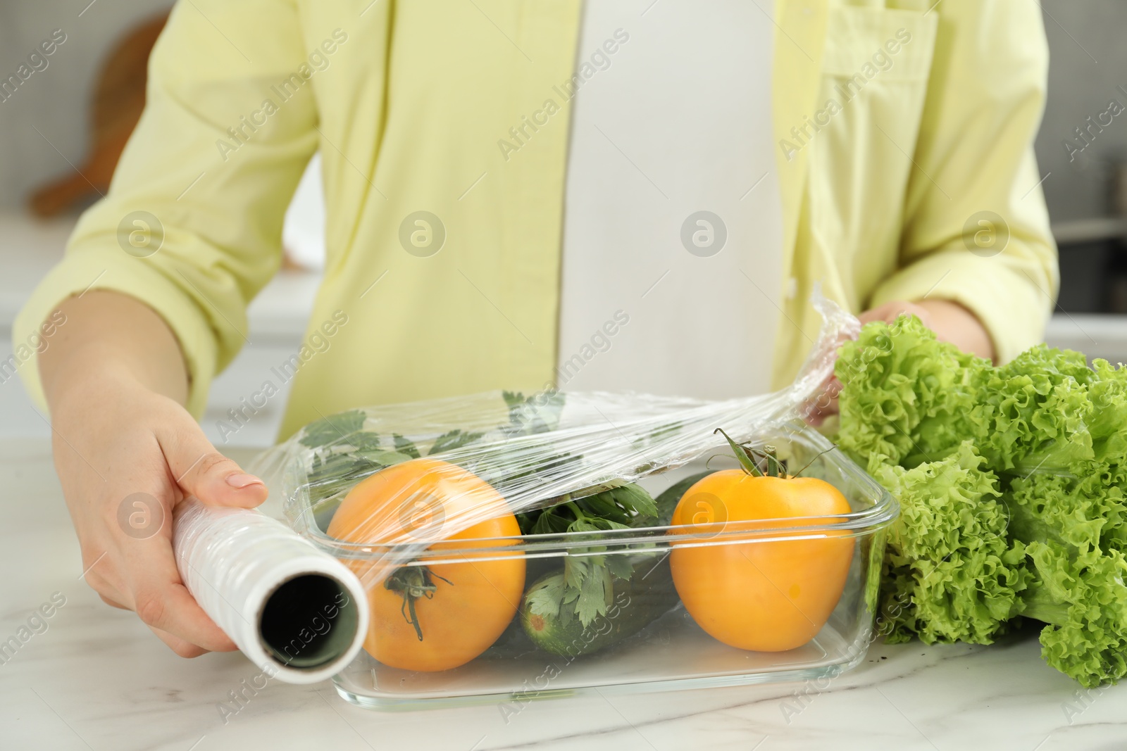 Photo of Woman putting plastic food wrap over glass container with fresh vegetables at white marble table, closeup