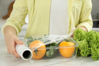 Woman putting plastic food wrap over glass container with fresh vegetables at white marble table, closeup