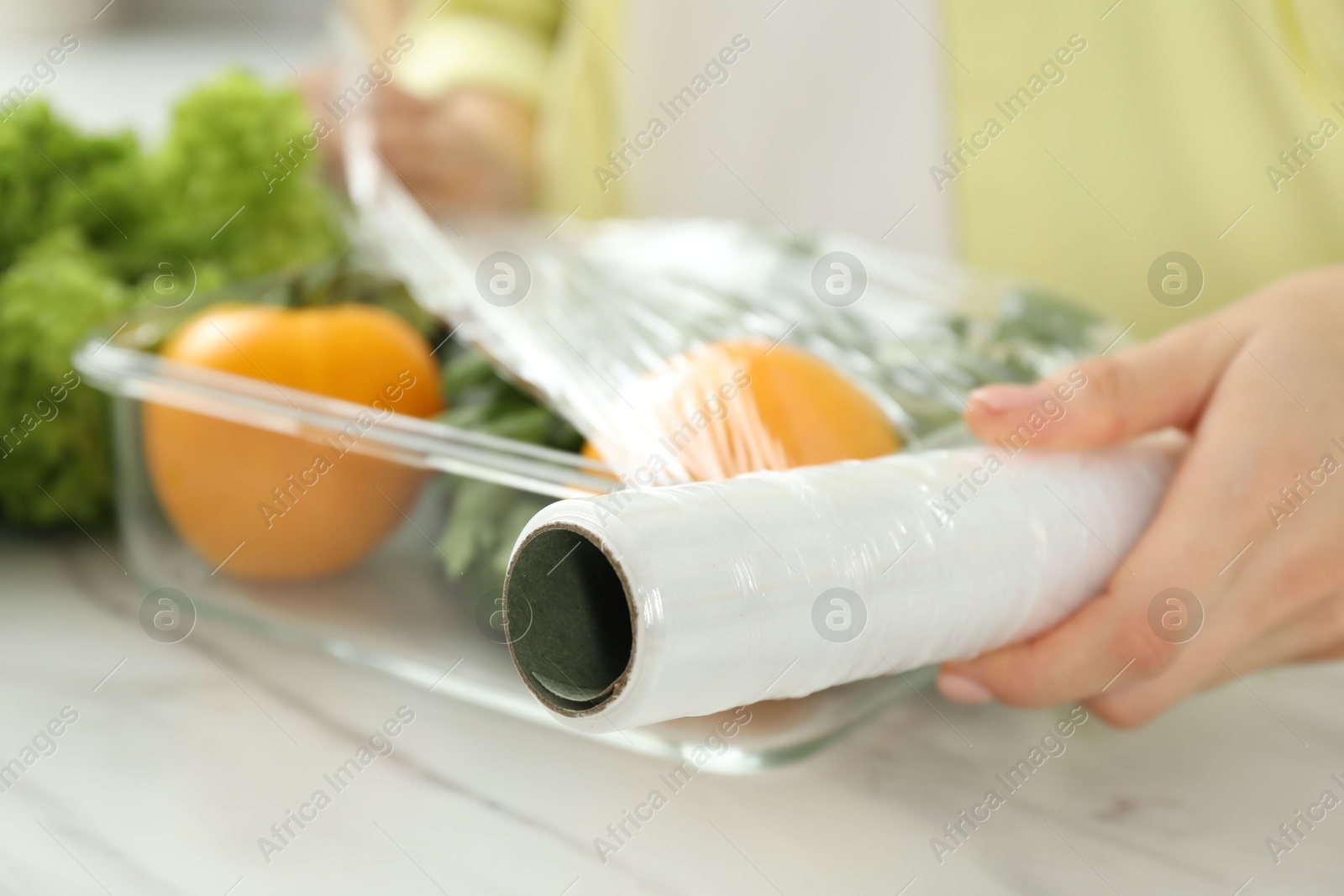 Photo of Woman putting plastic food wrap over glass container with fresh vegetables at white marble table, closeup