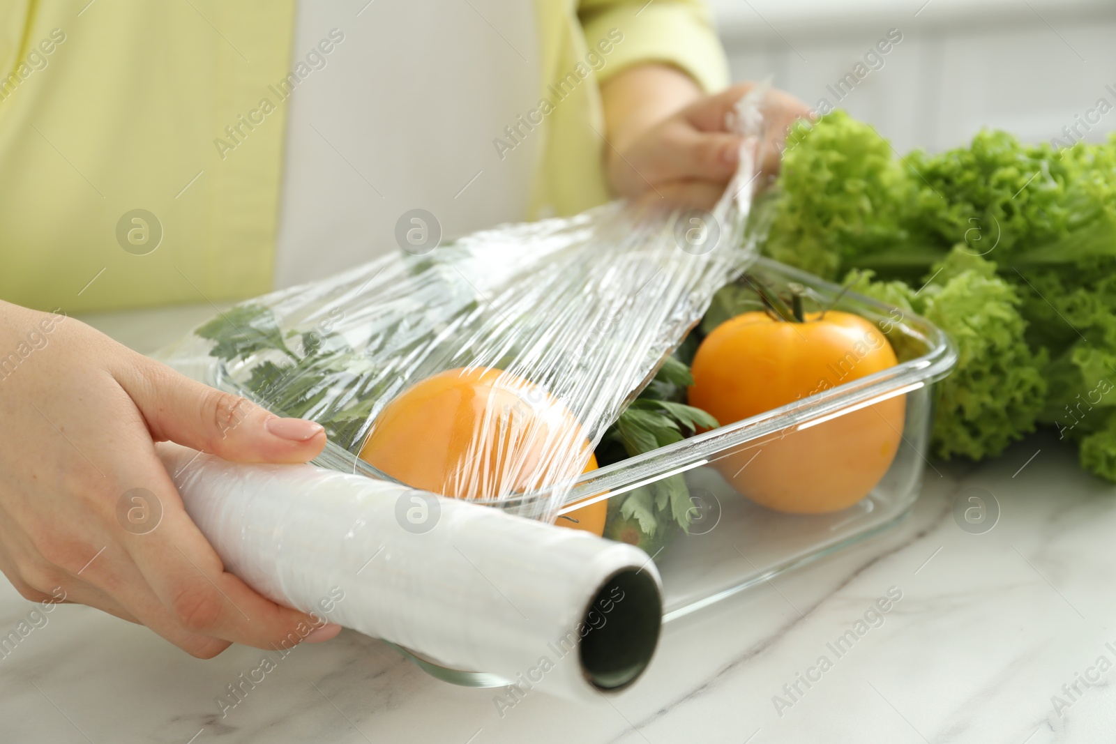 Photo of Woman putting plastic food wrap over glass container with fresh vegetables at white marble table, closeup