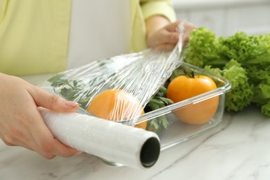 Woman putting plastic food wrap over glass container with fresh vegetables at white marble table, closeup