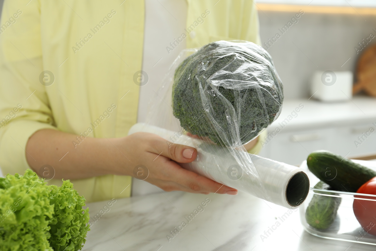 Photo of Woman putting plastic food wrap over broccoli at countertop in kitchen, closeup