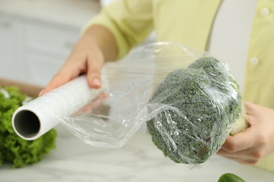 Woman putting plastic food wrap over broccoli at countertop in kitchen, closeup