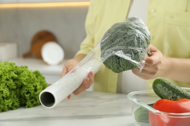 Photo of Woman putting plastic food wrap over broccoli at countertop in kitchen, closeup