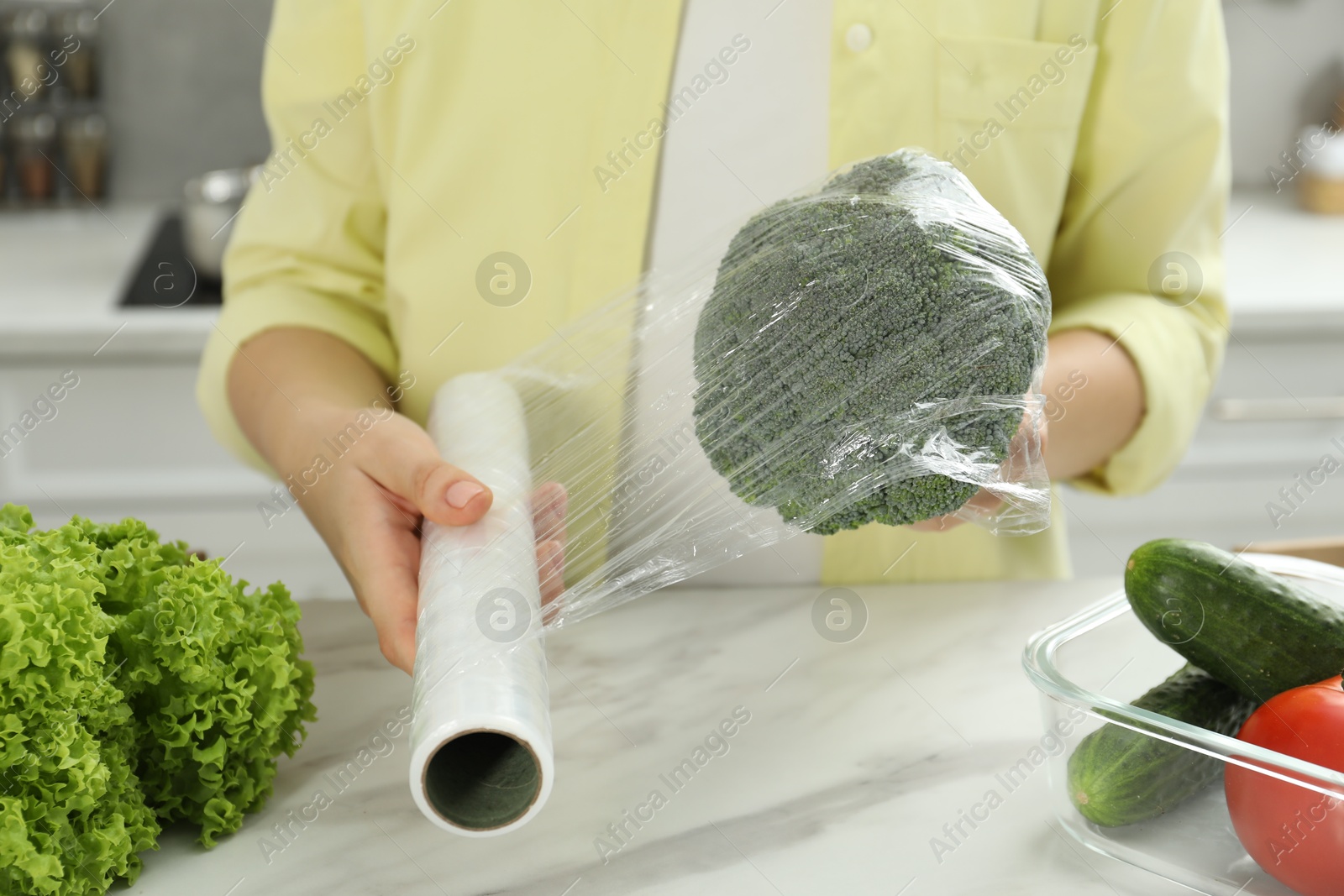 Photo of Woman putting plastic food wrap over broccoli at countertop in kitchen, closeup