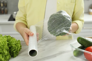 Woman putting plastic food wrap over broccoli at countertop in kitchen, closeup