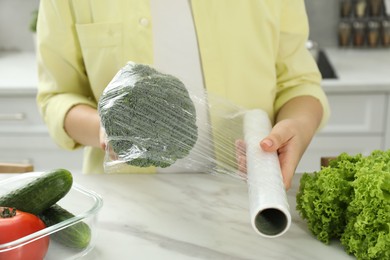 Woman putting plastic food wrap over broccoli at countertop in kitchen, closeup