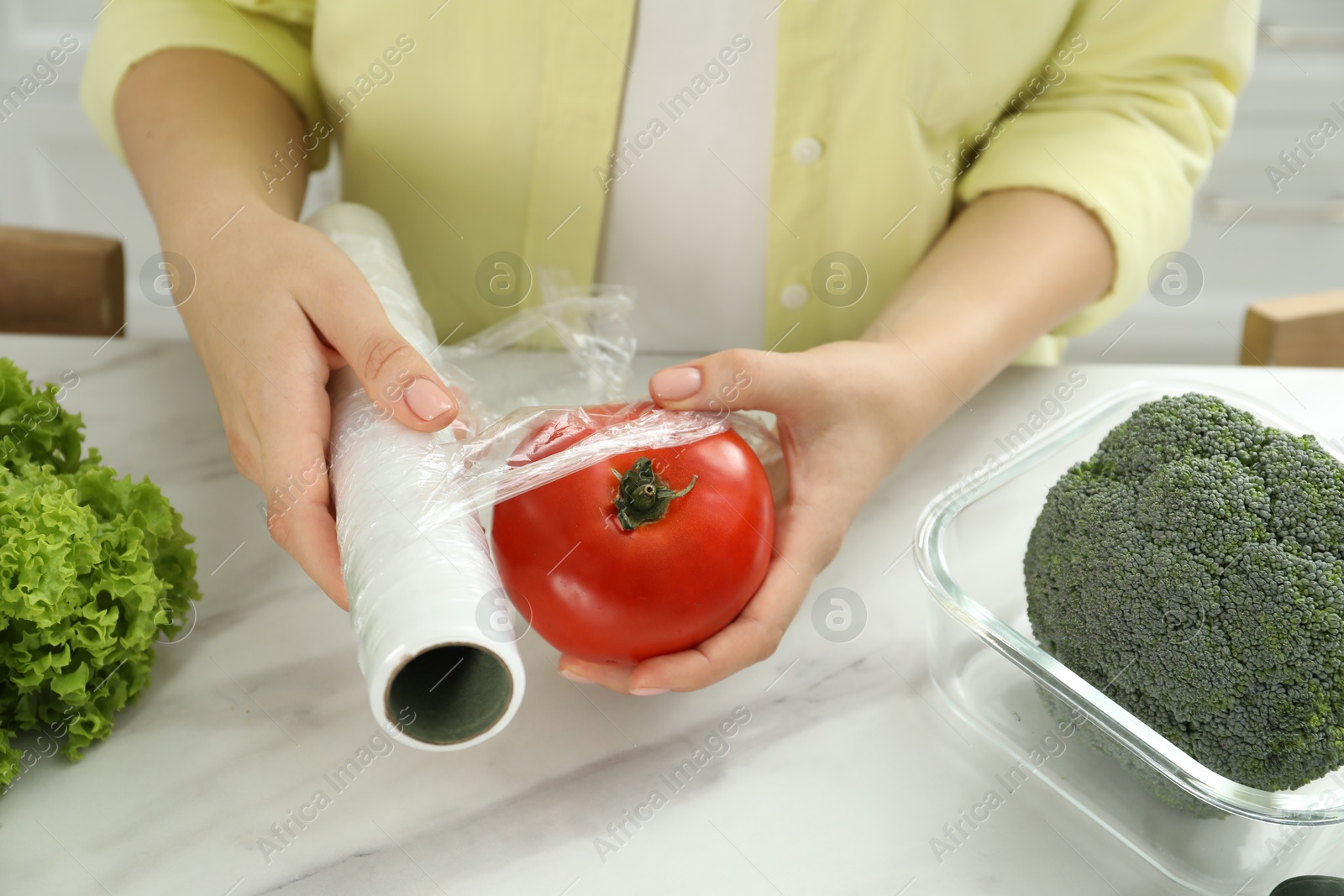 Photo of Woman putting plastic food wrap over tomato at countertop in kitchen, closeup