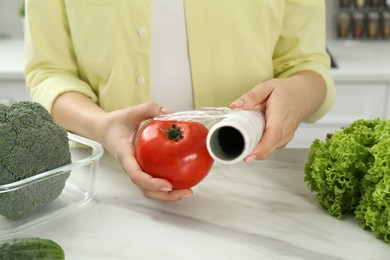 Woman putting plastic food wrap over tomato at countertop in kitchen, closeup