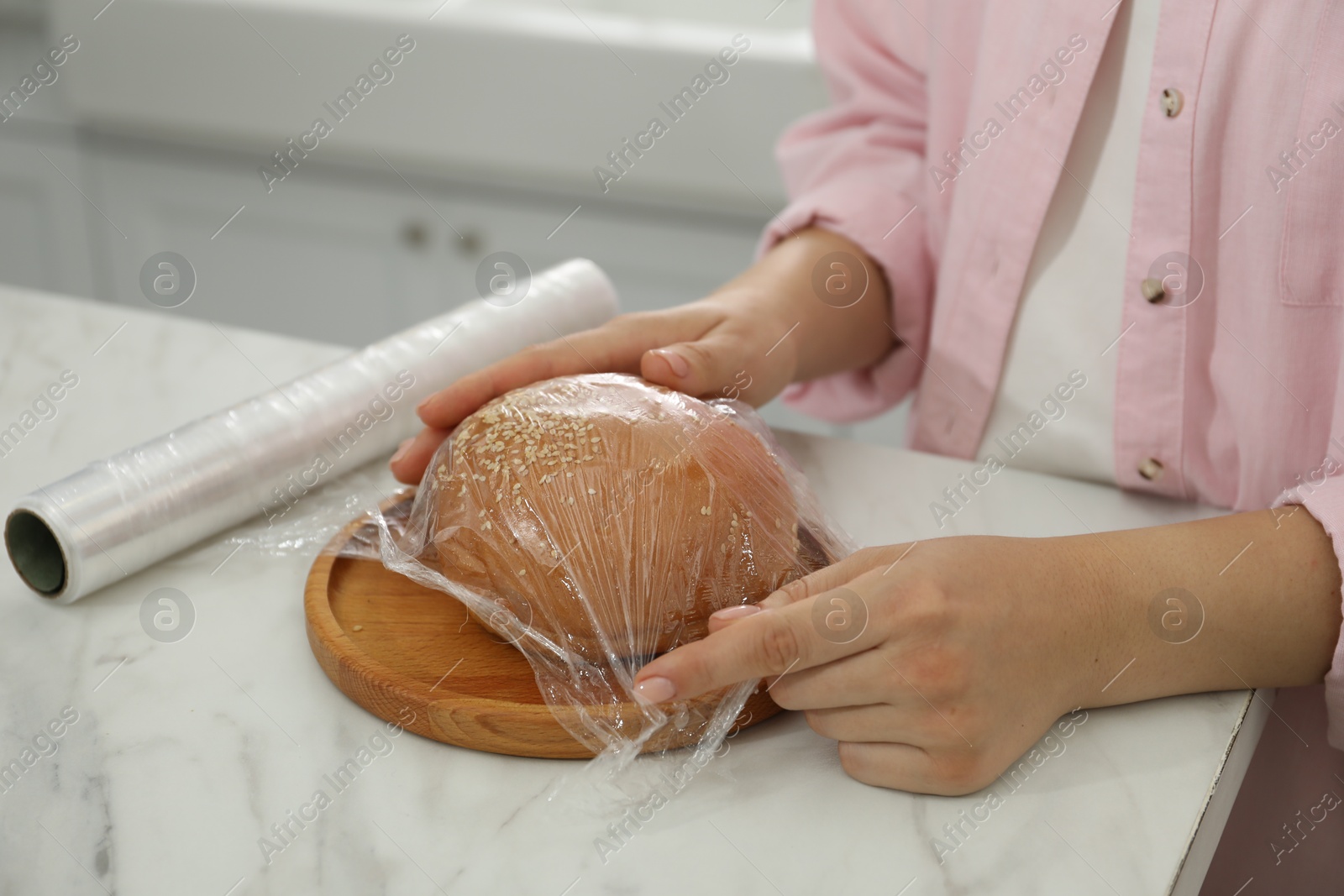 Photo of Woman putting plastic food wrap over fresh bun at white marble table in kitchen, closeup