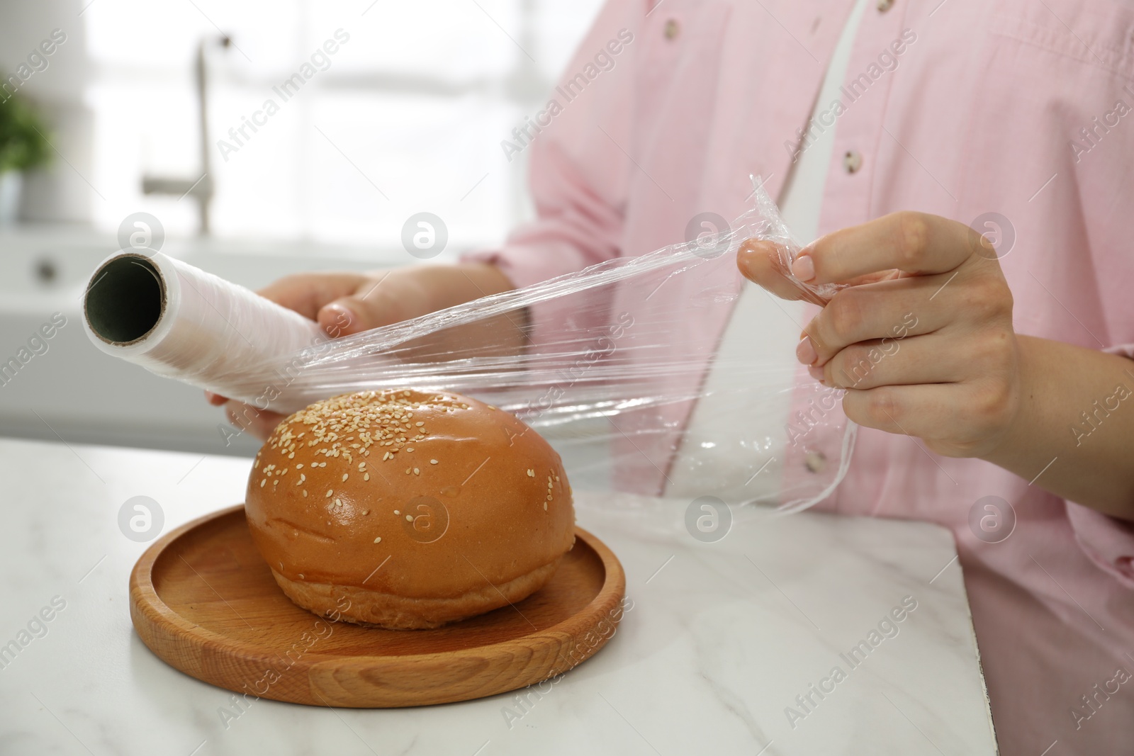 Photo of Woman putting plastic food wrap over fresh bun at white marble table in kitchen, closeup