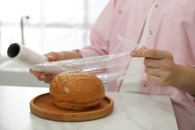 Photo of Woman putting plastic food wrap over fresh bun at white marble table in kitchen, closeup