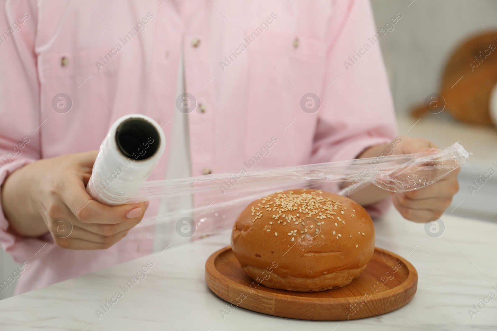 Photo of Woman putting plastic food wrap over fresh bun at white marble table in kitchen, closeup