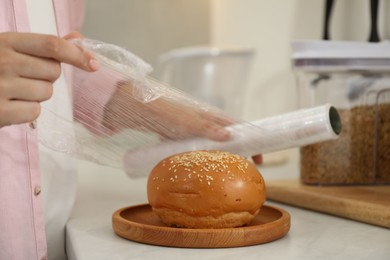 Photo of Woman putting plastic food wrap over fresh bun at white table in kitchen, closeup